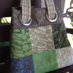 A patchwork tote bag with green, blue, and beige squares and two thick handles resting on a wooden bench.