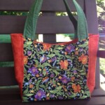 A floral-patterned tote bag with green handles and orange sides, resting on a wooden bench with greenery in the background.