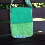 Green quilted shoulder bag with a patterned flap and strap, placed on a wooden bench with a natural background.