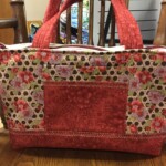 A red and floral-patterned quilted handbag is placed on a wooden table with a chair in the background.