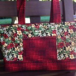 A red and floral-patterned handbag with two handles is placed on a wooden bench outdoors.