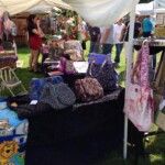 Outdoor craft fair with tables displaying colorful bags. Shoppers browse under white tents on a sunny day.