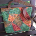 Patchwork quilted tote bag with green, orange, and red fabric patterns, resting on a wooden bench.