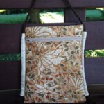 A floral-patterned fabric bag with a zipper pocket and rope handles hangs against a wooden surface.
