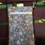 A fabric bag with a colorful circular pattern, featuring a zipper pocket, is displayed on a wooden bench.