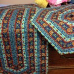 Quilt with geometric, multicolor pattern draped over a wooden surface. A pile of folded fabric is in the background.