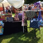 Outdoor market stall displaying colorful handmade bags and quilts under a tent, with a crowd walking by.