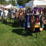 Outdoor craft fair with people browsing colorful bags displayed on stands under white tents. Grass and cloudy sky in the background.