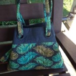 Handbag with green and gold feather pattern on a wooden bench with outdoor greenery in the background.