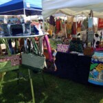 Outdoor market stall displaying colorful bags and textiles under white tents on grassy area.
