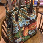 A floral and striped fabric tote bag with round handles hangs on a wooden chair. Background shows fabric rolls.