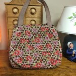 A floral-patterned handbag sits on a wooden table next to a lamp and a small figurine, with a wooden cabinet in the background.