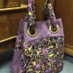 A floral-patterned fabric tote bag with purple accents and large gold eyelet handles, placed on a carpeted floor.