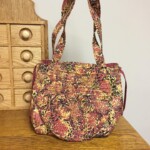 A patterned fabric handbag with floral design rests on a wooden table near a wooden drawer unit.
