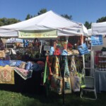 Craft booth at an outdoor market displaying colorful bags, quilts, and jars. Signs read "Creative Craftworks" and "10 Year Vendor.