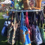 A display of colorful tote bags hanging on a metal rack at an outdoor market, with a booth and green grass in the background.