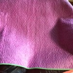 A pink quilt with a textured pattern and green border is draped over furniture near a window, with sunlight casting shadows.