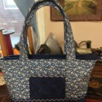 A blue and white patterned fabric tote bag with a pocket is displayed on a wooden table. A framed picture is in the background.
