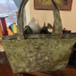 A green patterned tote bag with two handles, displayed on a wooden surface in a room with sewing thread spools in the background.