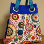 Colorful tote bag with patterned circles and vibrant blue handles, set on a wooden surface against a plain background.