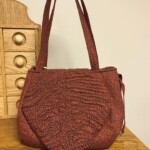 Red patterned handbag with two straps on a wooden surface next to a wooden cabinet.