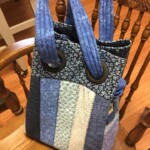 Patchwork blue fabric tote bag with two handles, featuring different patterned panels, resting on a wooden chair.