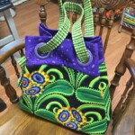 Colorful fabric bag with green and purple patterns, floral design, and green striped handles, placed on a wooden chair.