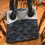 Blue and gray patterned tote bag with large black grommets and straps, resting on a wooden chair.