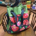 A colorful fabric tote bag with floral and striped patterns is placed on a wooden chair, surrounded by shelves of fabric.