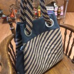 A striped tote bag with large metal eyelets and fabric handles rests on an ornate wooden chair.