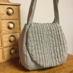 Gray textured handbag with vertical lines sits on a wooden surface next to a small wooden drawer unit.