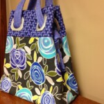 Colorful fabric tote bag with blue handles, featuring a floral pattern of blue roses and green leaves, resting on a wooden surface.