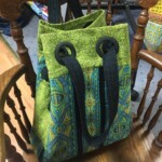 Green and blue patterned tote bag with black handles, resting on a wooden chair.