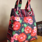 Quilted tote bag with red floral and striped patterns, featuring rings attaching the handles, placed on a wooden surface.