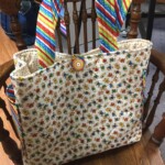 A colorful tote bag with striped handles and a button closure hangs on a wooden chair.
