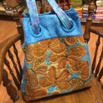 Patterned tote bag with blue and orange paisley design on a wooden chair, surrounded by fabric stacks.