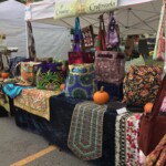Craft fair booth displaying colorful handmade bags and quilts on tables, with a sign reading "Creative Craftworks.