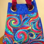 Colorful tote bag with swirling rainbow patterns and red handles, displayed on a wooden surface.