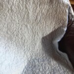 A quilt with abstract swirl patterns lies draped over a dark surface near a window.