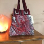 A handmade tote bag with red, black, and white abstract patterns sits on a wooden dresser next to a glowing orange lamp.