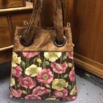 A floral patterned tote bag with braided handles is placed on a carpeted floor beside a wooden cabinet.