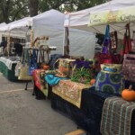 Outdoor craft market with booths featuring colorful quilts, bags, and pumpkins. Trees in the background.