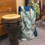 A djembe drum and a floral-patterned bag are placed on a carpeted floor next to a wooden dresser and a mirror.