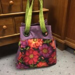 A floral-patterned handbag with green handles is placed on a carpeted floor near a wooden dresser.