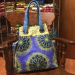 Colorful patterned bag with circular designs sits on a wooden chair, surrounded by neatly stacked fabric on metal shelves.