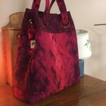 Red and purple patterned tote bag with handles, placed on a wooden table beside a decorative lamp.