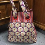 A multicolored tote bag with floral and striped patterns, featuring red circular handles, placed on a carpeted floor.