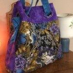 A colorful handbag with a floral pattern, featuring purple sides and blue handles, placed on a wooden surface next to a lamp.