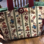 A fabric tote bag with a vintage-style pattern featuring dolls and toys, with red handles, placed on a wooden chair.