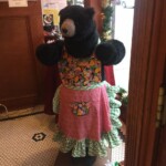 A bear statue dressed in a colorful floral apron stands inside a room near a decorated wall and door.